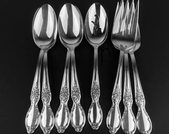 May include: A set of silver-colored flatware, including spoons and forks, arranged against a black background. The spoons have oval bowls and ornate floral detailing on the handles. The forks have four tines.