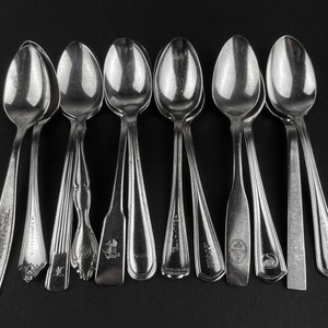 May include: A set of ten silver spoons with different engraved designs and brand names. The spoons are arranged in a row on a black surface.
