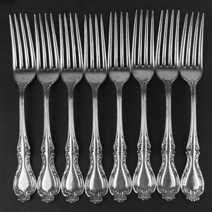 May include: Nine silver forks displayed in a row. Each fork features a decorative handle with an ornate floral design. The tines are long and slender. The forks are set against a dark background.