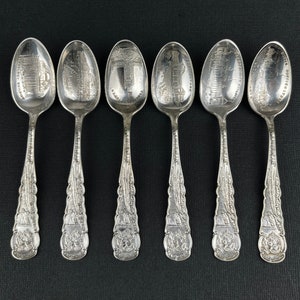 May include: Six silver spoons with detailed engravings of buildings and landmarks. Each spoon features a different design on the bowl, with text and ornate handles. The spoons are arranged on a black surface.
