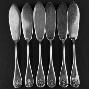 May include: Six silver butter knives with ornate handles arranged in a row against a black background.