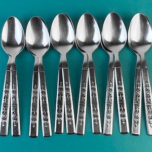 May include: Seven stainless steel spoons arranged on a teal surface. Each spoon features a rounded bowl and a handle adorned with floral detailing. The spoons reflect the surrounding environment. The spoons are made of stainless steel.