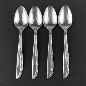 May include: Four silver teaspoons with ornate floral designs on the handles. The spoons are arranged in a row on a black surface.