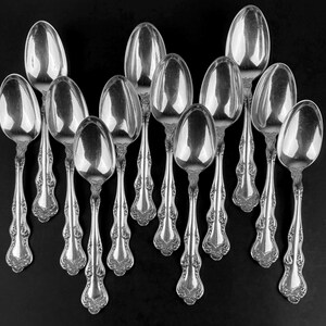 May include: An arrangement of silver spoons with decorative handles on a black background. The spoons have an ornate design with floral patterns and oval bowls. The reflective surfaces of the spoons create highlights and shadows.