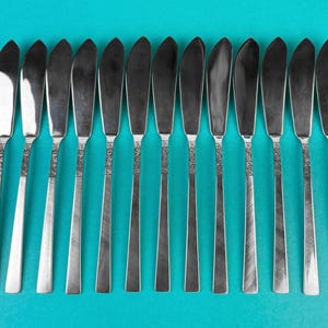 May include: A set of 14 silver butter knives with textured handles. The knives are arranged in a row on a blue background.