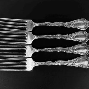 May include: Four antique silver forks with ornate handles and tines, arranged horizontally against a black background. Each fork features a decorative design on the handle and a personalized inscription. The forks are a classic example of vintage silverware.
