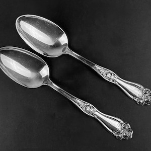 May include: Two silver spoons with ornate rose designs on the handles. The spoons are polished and reflect light.