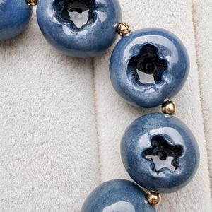 Könnte beinhalten: Ein Armband mit mehreren kugelförmigen, heidelbeerförmigen blauen Keramikperlen, die jeweils eine dunkle, sternförmige Vertiefung in der Mitte aufweisen. Die blauen Perlen sind durch kleine, polierte goldfarbene Metallzwischenperlen getrennt.