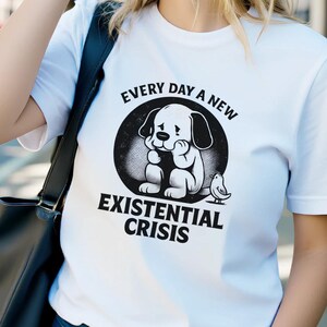 Könnte beinhalten: Weißes T-Shirt mit dem Text "EVERY DAY A NEW EXISTENTIAL CRISIS" und einer Grafik eines traurigen Hundes. Das T-Shirt wird im Freien getragen. Die Person trägt eine Sonnenbrille und blaue Jeans.
