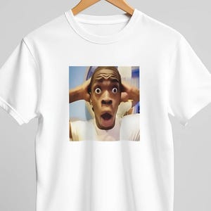 Könnte beinhalten: Weißes T-Shirt mit einem Meme einer überraschten Person, die sich die Hände an den Kopf hält und den Mund geöffnet hat. Das Bild ist quadratisch und auf dem T-Shirt zentriert. Das T-Shirt hängt an einem Holzbügel.