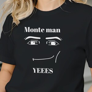 Può includere: T-shirt nera con la scritta "Monte man" sopra una grafica stilizzata del volto e la parola "YEEES" sotto. La grafica presenta un paio di occhi e una bocca sorridente. La maglietta è realizzata in materiale morbido.