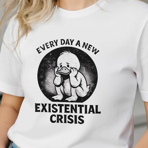 Könnte beinhalten: Weißes T-Shirt mit einem schwarzen Aufdruck einer Ente, die ihren Kopf in den Händen hält, und dem Text "EVERY DAY A NEW EXISTENTIAL CRISIS" in einem kreisförmigen Design. Das T-Shirt wird getragen.