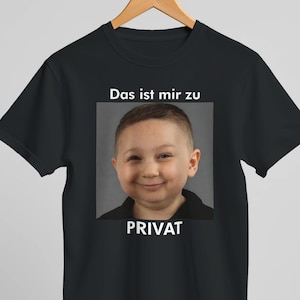 Könnte beinhalten: Schwarzes T-Shirt mit dem lächelnden Gesicht eines Kindes in einem quadratischen Rahmen. Der Text über dem Bild lautet "Das ist mir zu", und unter dem Bild steht "PRIVAT". Das T-Shirt hängt an einem Holzbügel.