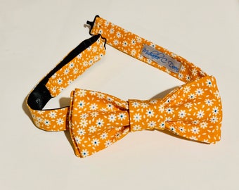 May include: A yellow bow tie with a white daisy pattern and blue centers. The bow tie is adjustable and has a black interior. The tag reads "Michelle C. Deer".