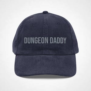 May include: A navy blue corduroy baseball cap featuring the text DUNGEON DADDY embroidered in light grey across the front panel. The hat has a structured crown, a curved brim, and a button at the top, showcasing a classic six-panel design.