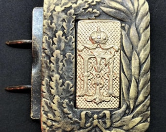 Puede incluir: Una hebilla de cinturón de metal dorado con un diseño ornamentado. La hebilla presenta una corona, las letras "F.I." y el número "63".