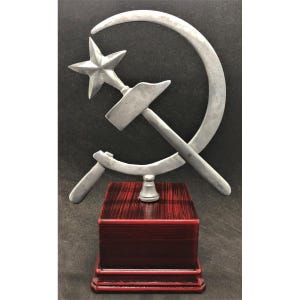 Könnte beinhalten: Eine silberne Metallskulptur eines Hammers und einer Sichel auf einem roten Holzsockel. Hammer und Sichel sind ein Symbol des Kommunismus.