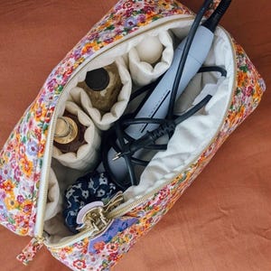 Op de afbeelding: Make-uptas met bloemenprint en een gouden rits, open om compartimenten te tonen met schoonheidsproducten en een grijze haarstylingtool. De binnenkant van de tas is wit en de buitenkant heeft een kleurrijk bloemenpatroon.