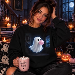 Peut inclure: Un sweat-shirt noir à col rond orné d'un motif représentant un fantôme blanc soufflant une bulle de chewing-gum rose. Le sweat-shirt est accompagné d'une tasse en céramique rose clair avec le texte Spooky Vibes et un petit cœur imprimés.
