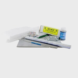 May include: A glass beadmaking kit including a Hot Head torch in a yellow and white box, a blue bottle of Air-Dri, a grey work surface, a stack of white ceramic fiber insulation, metal mandrels, and a small bag of assorted glass beads.