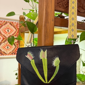 May include: Black leather crossbody bag featuring a hand-painted design of Venus flytraps in green, pink, and white. The bag has a flap closure and a yellow and gray patterned strap. The bag is hanging in front of a wooden background.