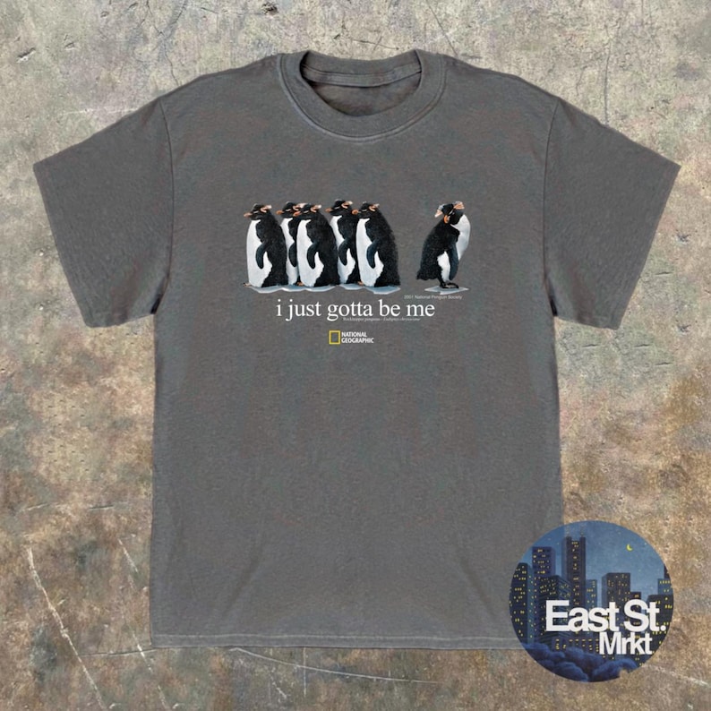 Może przedstawiać: Ciemnoszary t-shirt z kr&oacute;tkim rękawem z grafiką przedstawiającą pięć pingwin&oacute;w w rzędzie, z napisem "i just gotta be me" oraz logo National Geographic nadrukowanym na biało na środku klatki piersiowej.