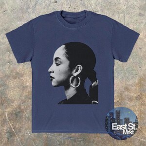 May include: Navy blue t-shirt featuring a black and white portrait of a woman with hoop earrings. The image is a side profile. A circular logo with the text "East St. Mrkt" is on the bottom right.