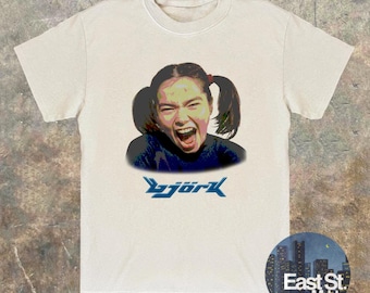May include: Cream-colored t-shirt featuring a graphic of a person with pigtails, screaming with their mouth open. The word "Björk" is printed in blue, stylized text below the image. A circular logo with the text "East St. Mrkt" is in the bottom right corner.