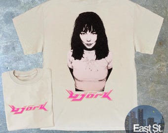 May include: A beige t-shirt with a pink and white graphic of Bjork, a singer, with the word "Bjork" in pink letters below the image.
