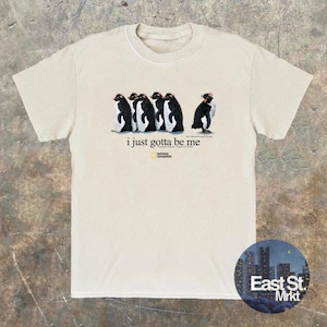 Może przedstawiać: Kremowy t-shirt z kr&oacute;tkim rękawem z grafiką przedstawiającą pięć pingwin&oacute;w w rzędzie, z napisem "i just gotta be me" oraz logo National Geographic nadrukowanym pod pingwinami na środku klatki piersiowej.