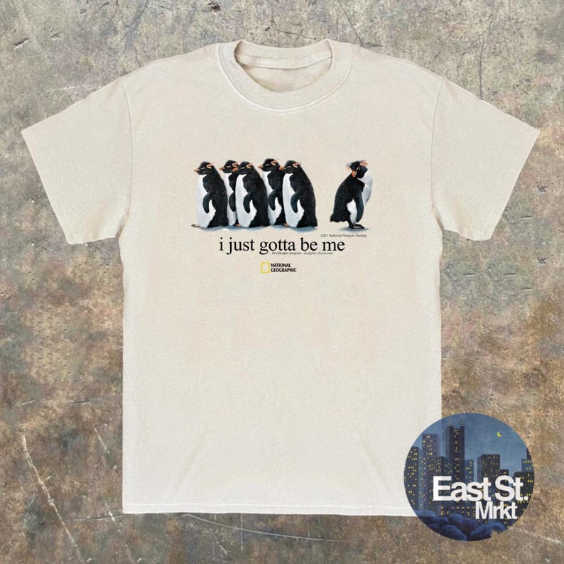 Może przedstawiać: Kremowy t-shirt z kr&oacute;tkim rękawem z grafiką przedstawiającą pięć pingwin&oacute;w w rzędzie, z napisem "i just gotta be me" oraz logo National Geographic nadrukowanym pod pingwinami na środku klatki piersiowej.