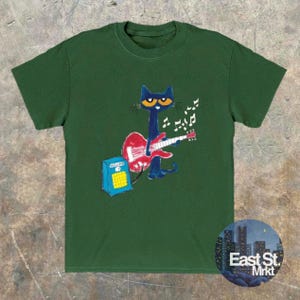 May include: Dark green t-shirt with a cartoon cat playing a red guitar, accompanied by musical notes. The cat is blue with yellow eyes. A blue amplifier is also present. The shirt features the text "East St. Mrkt" in a circular design.
