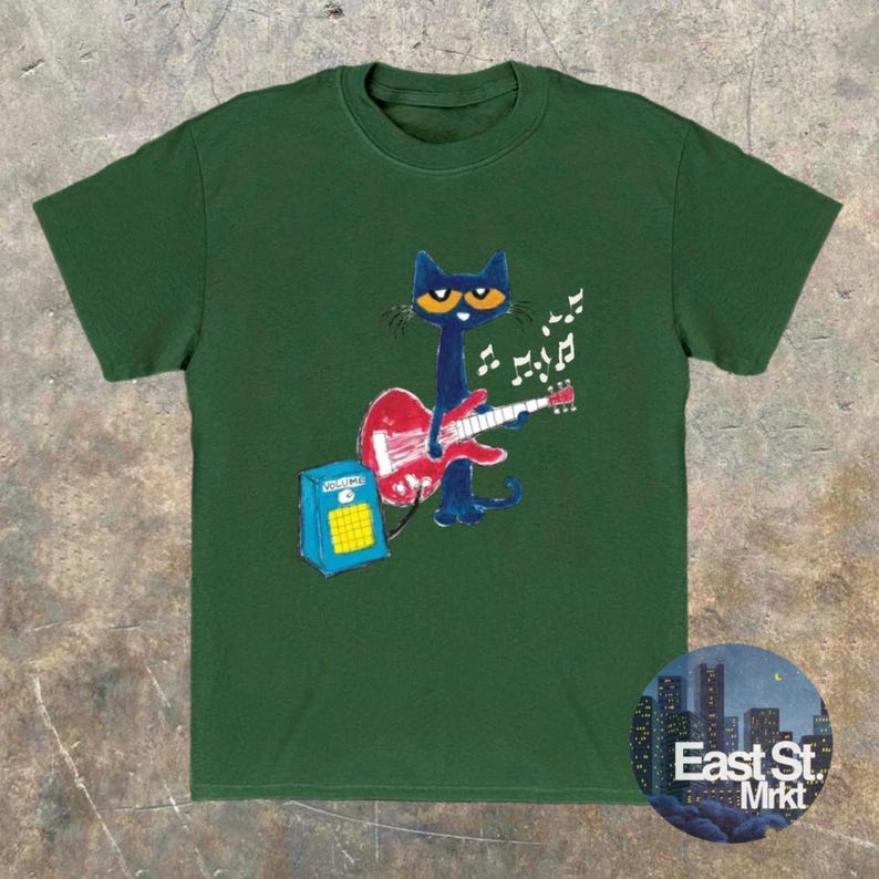 May include: Dark green t-shirt with a cartoon cat playing a red guitar, accompanied by musical notes. The cat is blue with yellow eyes. A blue amplifier is also present. The shirt features the text "East St. Mrkt" in a circular design.