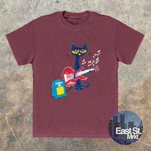 May include: Burgundy t-shirt with a cartoon cat playing a red guitar, accompanied by musical notes. The cat is black with yellow eyes. A blue amplifier is next to the cat. The shirt has a round neck and short sleeves. The East St. Mrkt logo is in the bottom right corner.