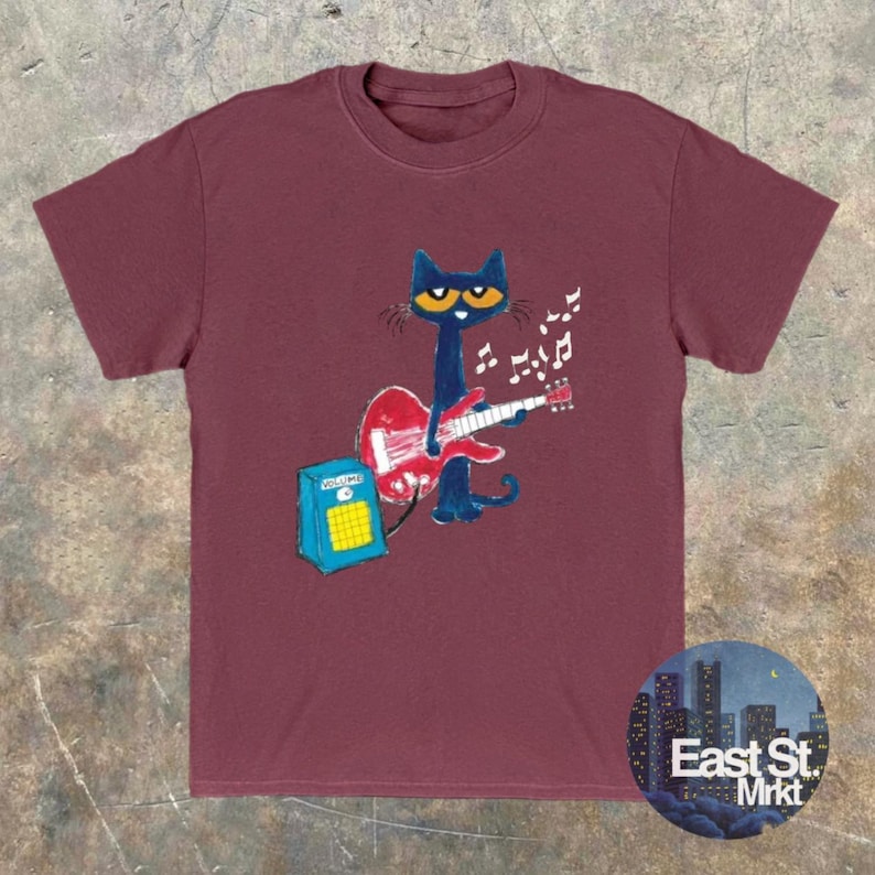 May include: Burgundy t-shirt with a cartoon cat playing a red guitar, accompanied by musical notes. The cat is black with yellow eyes. A blue amplifier is next to the cat. The shirt has a round neck and short sleeves. The East St. Mrkt logo is in the bottom right corner.