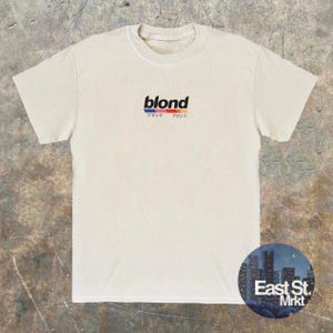 Puede incluir: Una camiseta blanca con la palabra "blond" impresa en letras negras con una franja roja, azul y amarilla debajo. Las palabras japonesas "ブラック" y "ブロンド" están impresas debajo de la palabra "blond".