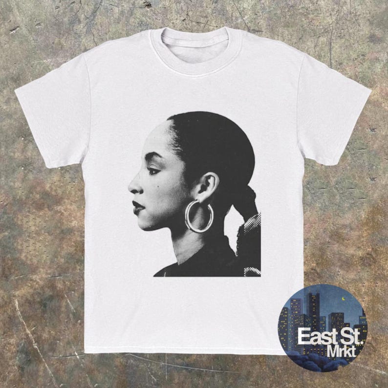 May include: White t-shirt featuring a black and white profile portrait of a woman with hoop earrings. The image is a side view of the woman's face and hair. A circular logo with the text "East St. Mrkt" is in the lower right corner.