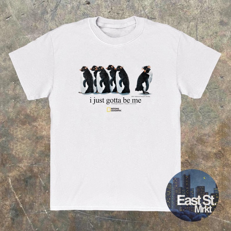 Może przedstawiać: Biały t-shirt z kr&oacute;tkim rękawem z grafiką przedstawiającą pięć pingwin&oacute;w idących w rzędzie, z kt&oacute;rych jeden jest zwr&oacute;cony w przeciwnym kierunku. Tekst brzmi i just gotta be me i zawiera logo National Geographic pod pingwinami.