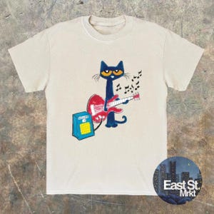 May include: Beige t-shirt with a cartoon design of a blue cat playing a red guitar, with musical notes above. A blue amplifier is to the left. The shirt has a round neck and short sleeves. The words "East St. Mrkt" are in a circle at the bottom.