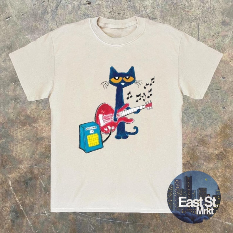 May include: Beige t-shirt with a cartoon design of a blue cat playing a red guitar, with musical notes above. A blue amplifier is to the left. The shirt has a round neck and short sleeves. The words "East St. Mrkt" are in a circle at the bottom.