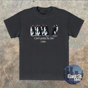 Może przedstawiać: Czarny t-shirt z kr&oacute;tkim rękawem z grafiką przedstawiającą pięć pingwin&oacute;w stojących w rzędzie, z kt&oacute;rych jeden patrzy w przeciwnym kierunku. Tekst "i just gotta be me" znajduje się nad ż&oacute;łto-czarnym logo National Geographic.