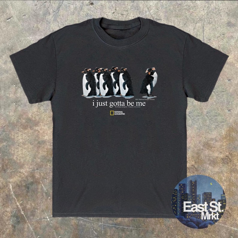 Może przedstawiać: Czarny t-shirt z kr&oacute;tkim rękawem z grafiką przedstawiającą pięć pingwin&oacute;w stojących w rzędzie, z kt&oacute;rych jeden patrzy w przeciwnym kierunku. Tekst "i just gotta be me" znajduje się nad ż&oacute;łto-czarnym logo National Geographic.