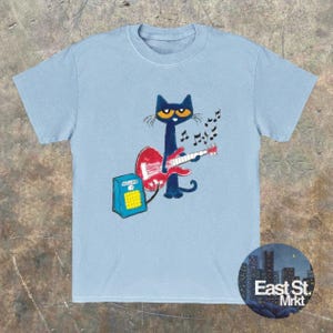 May include: Pale blue t-shirt with a cartoon cat playing a red guitar. The cat has yellow eyes and is in front of a blue amplifier. Musical notes are above the guitar. The shirt has a round neck.