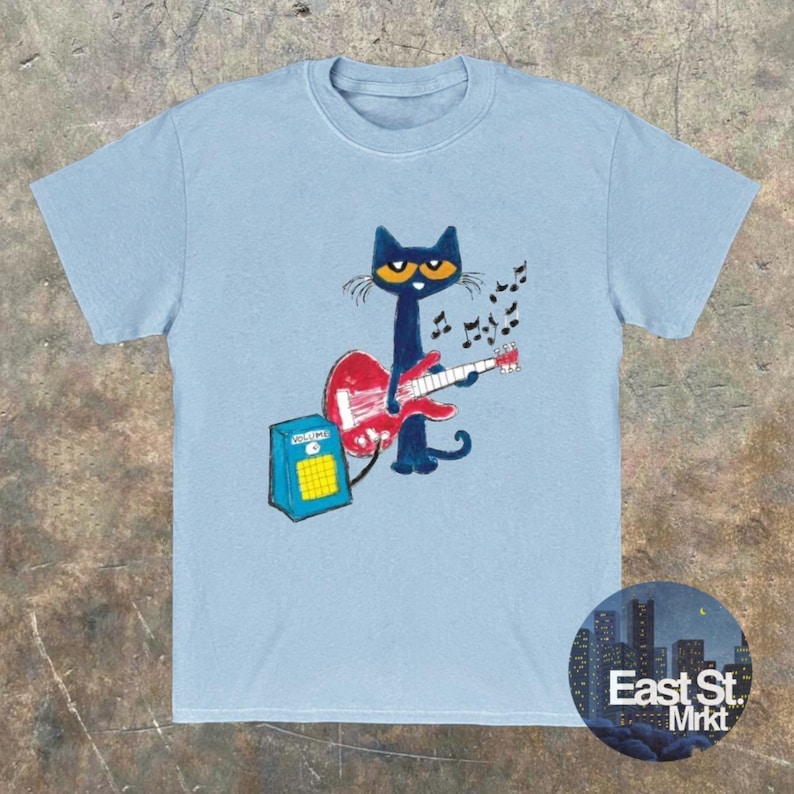May include: Pale blue t-shirt with a cartoon cat playing a red guitar. The cat has yellow eyes and is in front of a blue amplifier. Musical notes are above the guitar. The shirt has a round neck.