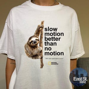 May include: A white t-shirt featuring a graphic of a brown sloth hanging from a tree branch. The text reads "slow motion better than no motion" followed by "2001 sloth appreciation society" and the National Geographic logo in yellow and black.