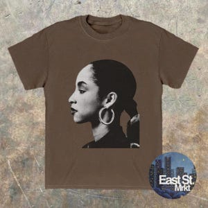 May include: Brown t-shirt featuring a black and white profile portrait of a woman with hoop earrings. The image is a graphic design. The shirt has a crew neck and short sleeves. A circular logo with the text "East St. Mrkt" is on the bottom right.