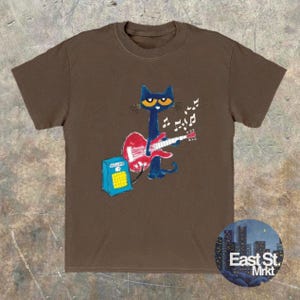 May include: A brown t-shirt with a cartoon cat playing a red guitar, with musical notes floating above. The cat is blue with yellow eyes. A blue amplifier is next to the cat. The shirt has the text "East St. Mrkt" on the bottom right.