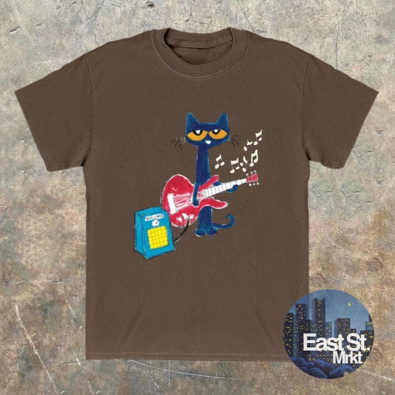 May include: A brown t-shirt with a cartoon cat playing a red guitar, with musical notes floating above. The cat is blue with yellow eyes. A blue amplifier is next to the cat. The shirt has the text "East St. Mrkt" on the bottom right.