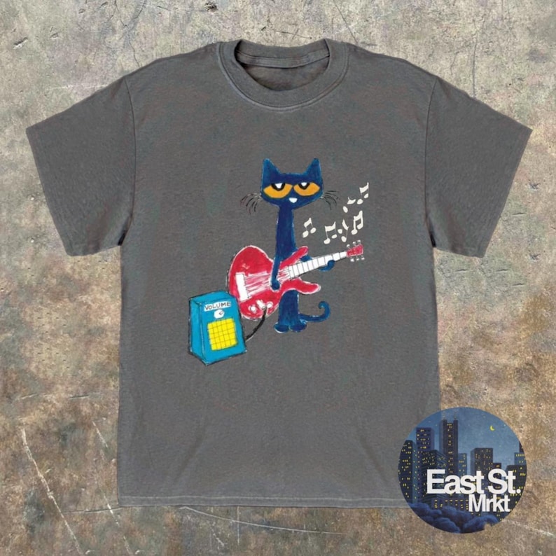 May include: A grey t-shirt with a cartoon cat playing a red guitar, with musical notes above. The cat is blue with yellow eyes. A blue amplifier is next to the cat. The shirt has a round neck and short sleeves.