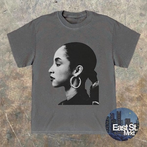 May include: Gray t-shirt featuring a black and white profile portrait of a woman with hoop earrings. The shirt has a crew neck and short sleeves. A circular logo with the text "East St. Mrkt" is in the lower right corner.